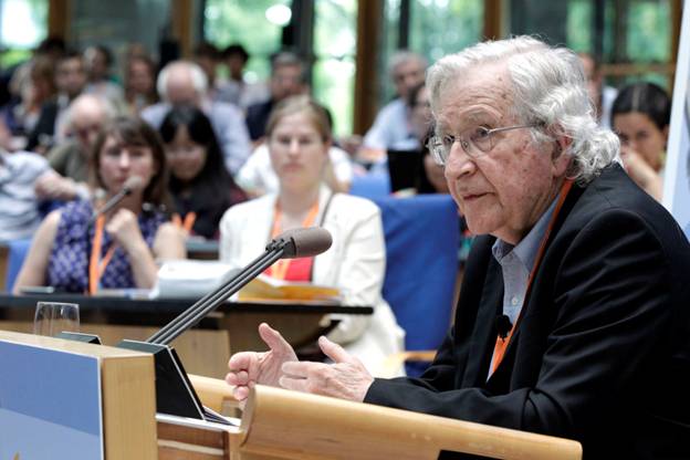 Noam Chomsky speaks while seated at a desk